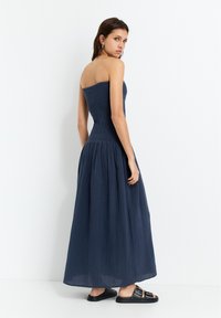 Marineblaues trägerloses Kleid mit gerafftem Oberteil und fließendem Rock, gefertigt aus strukturiertem Stoff. Kombiniert mit schwarzen Sandalen, vor einem weißen Hintergrund präsentiert.