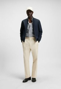 Navy bomber jacket, striped button-up shirt, beige trousers, black shoes, and a light-colored cap. Smooth fabric, tailored fit, modern design.