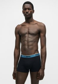 Homme athlétique à la peau foncée portant un boxer noir avec une ceinture bleue portant l'inscription "BOSS", debout devant un fond blanc uni.