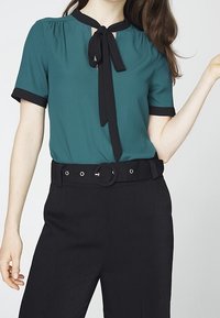 Blusa teal con maniche corte, rifiniture nere e un fiocco al collo, abbinata a pantaloni neri a vita alta con una grande fibbia circolare.