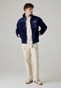 Jauns vīrietis, ģērbts tumši zilajā flīsa jakā, krēmkrāsas šortos un baltos sporta apavos, stāv ar rokām kabatās, skatoties pa labi.