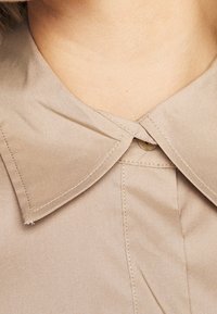 Beige skjorta med krage som har en slät textur, dubbelstitchade kanter och en central knappdetalj under kragen.