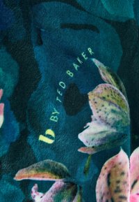 Bavlnená látka s kvetinovým vzorom v odtieňoch zelenej a modrej, s veľkými ružovými a fialovými kvetmi. Text znie "b by TED BAKER." Mäkká textura.