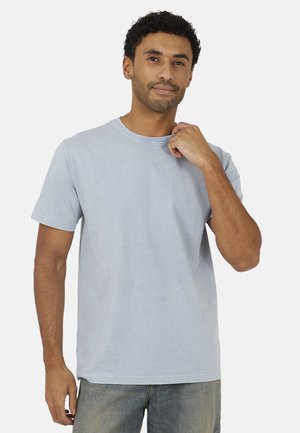 Uomo che indossa una maglietta a maniche corte azzurro chiaro e jeans, toccando il colletto, in piedi davanti a uno sfondo bianco uniforme.