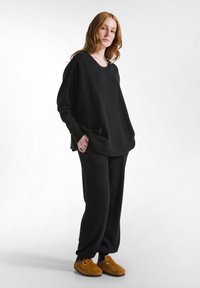 Giovane donna con capelli rossi che indossa un ampio maglione nero, pantaloni neri e pantofole marroni, in piedi su uno sfondo bianco.