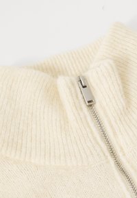 Close-up of a cream-colored ribbed knit sweater collar with a silver metal zipper partially zipped up.