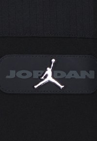Črn material z teksturiranim dizajnom, s poudarjenim srebrnim logotipom "Jordan" in ikonično silhueto Jumpman na usnjeni oznaki.