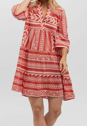 Frau trägt ein locker sitzendes, knielanges rotes Kleid mit weißen geometrischen Mustern und Dreiviertelärmeln vor einfarbigem Hintergrund.
