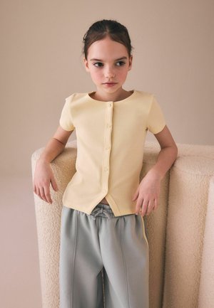 Jeune fille aux cheveux foncés portant un haut jaune pâle boutonné et un pantalon gris ample, debout contre un siège et un mur beige texturé.