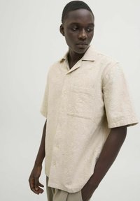 Camicia beige a maniche corte con un motivo floreale texturizzato, tasca frontale, chiusura a bottoni e una vestibilità rilassata. Tessuto morbido con una leggera lucentezza.