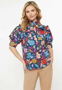 Bluse mit Blumenmuster und Puffärmeln, mit gebundenem Ausschnitt und blauem Hintergrund. Sie enthält lebendige Farben wie Rot, Pink und Gelb.