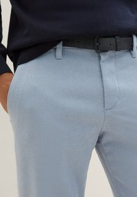 Man wearing light blue textured trousers with black belt and navy sweater, hand in left pocket, waist to mid-thigh view.