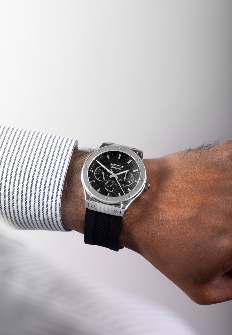 Montre analogique noire avec un cadran noir texturé, boîtier en métal argenté et bracelet en caoutchouc ; elle possède des sous-cadrans chronographes et des accents en acier inoxydable.