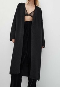 Black, long cardigan in soft, textured fabric with open front, relaxed fit, and dropped shoulders. Worn over matching black trousers and lace bra.