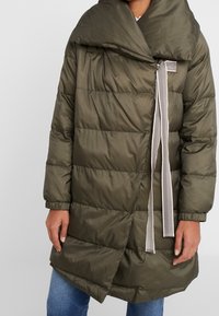 Vadderad kappa i olivgrönt med bred krage, asymmetriskt stängningssystem och ett randigt knytband som detalj. Slät, glansig yta med quiltad design.