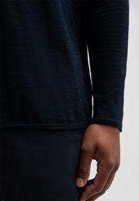 Langärmliges, dunkelblaues Hemd aus weichem, strukturiertem Stoff mit abgerundetem Saum und gestickten Akzenten, kombiniert mit enganliegenden, dunklen Hosen und einer Hand, die daneben ruht.