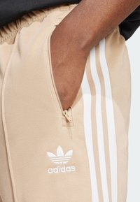Beige Adidas sweatpants with white three-stripe design, nylon material, side pocket with zipper, and embroidered logo on the thigh.