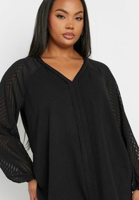 Black blouse with a V-neckline, long puff sleeves made of sheer textured fabric, and a loose fit. Features subtle geometric patterns on sleeves.