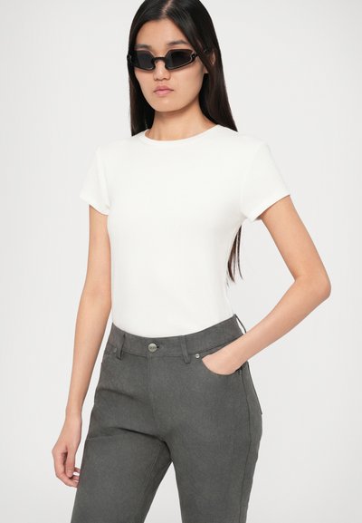 Γυναίκα με μακριά μαύρα μαλλιά, φορώντας μαύρα κομψά γυαλιά ηλίου, απλή λευκή μπλούζα κοντομάνικη και σκούρους γκρι παντελόνες με το ένα χέρι στη τσέπη.