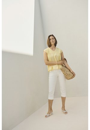 Gul bluse med pliser, hvide capribukser og lyse sandaler. Holder en vævet taske og står mod en ensfarvet, neutral baggrund.