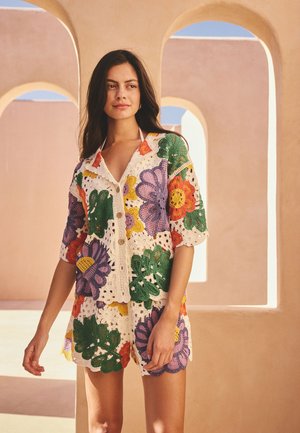 Femme aux longs cheveux foncés portant une chemise boutonnée en crochet et un short à grands motifs floraux colorés, debout près d'arcs de couleur pêche.