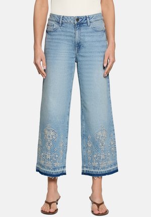 Jean bleu clair à jambes larges avec broderies florales blanches près des ourlets effilochés, porté avec des sandales marron en forme de tongs et un haut blanc côtelé.