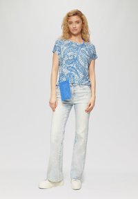 Blauw patroond blouse met korte mouwen, gecombineerd met lichte denim flared jeans en witte platform sneakers. Een blauwe gestructureerde telefoontas hangt van de schouder.
