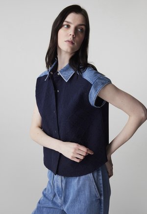 Vrouw die een mouwloze marineblauwe gewatteerde top draagt met een denim kraag en mouwen, gecombineerd met loszittende lichtblauwe denim broek, poseert zijwaarts.