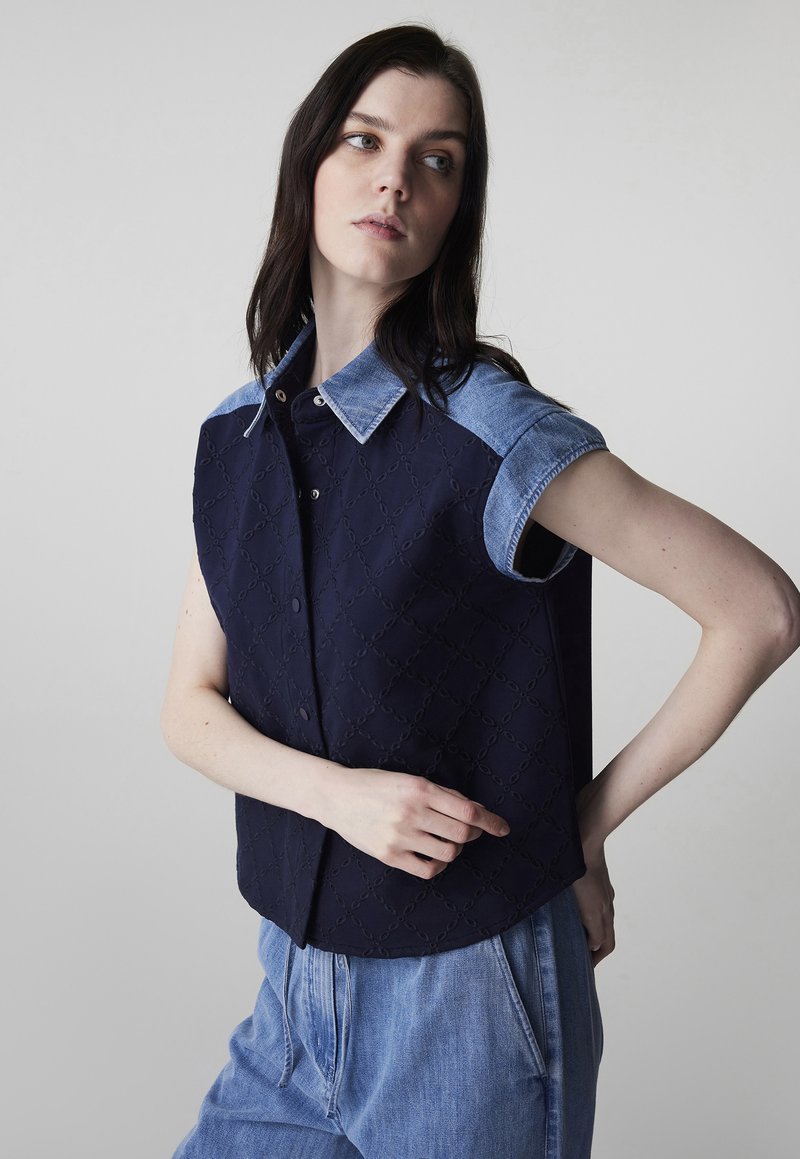 Frau trägt ein ärmelloses marineblaues, gesteppte Oberteil mit Jeanskragen und Ärmeln, kombiniert mit locker sitzenden hellblauen Jeanshosen, posiert seitlich.