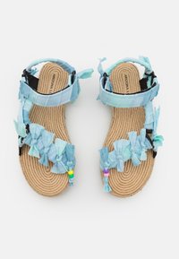 Barnsandaler med flätade bruna sulor och blå tygremmar prydda med rosetter och färgglada pärlor, sedda ovanifrån mot en vit bakgrund.