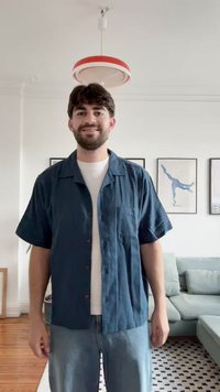 Camicia a maniche corte, con bottoni, in tessuto blu scuro con una vestibilità comoda, dotata di taschino sul petto e bottoni marrone chiaro, abbinata a jeans chiari.
