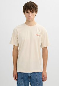 Joven con cabello castaño que lleva una camiseta beige clara con un pequeño texto rojo que dice "Yosemite" y jeans de mezclilla azul, sobre un fondo neutro.