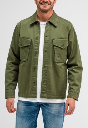 Olive green cotton jacket with a button-up front, two chest pockets, and a relaxed fit, layered over a white shirt. Denim jeans visible below.