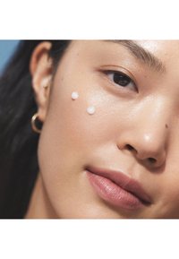 Gros plan d'un visage avec une peau lisse et lumineuse et deux petites gouttes de crème sur la joue. Boucle d'oreille visible dans l'oreille.