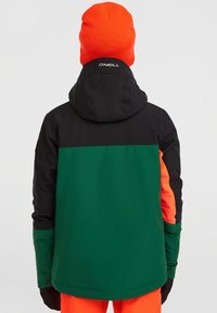 Färgglad skidjacka med svarta, gröna och orange paneler, huva och ergonomiska ärmar. Materialet ser strukturerat och vattentåligt ut.
