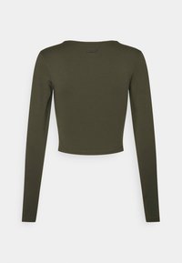 Długim rękawem krótkiego topu w kolorze oliwkowej zieleni, wykonanym z gładkiej, elastycznej tkaniny. Posiada okrągły dekolt i minimalne przeszycia. Logo nadrukowane na plecach.