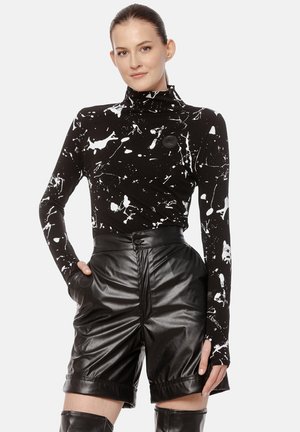 Kobieta nosząca czarną i białą bluzkę w splatery, wysokie skórzane szorty i czarne kozaki do kolan, pozująca z jedną ręką uniesioną.