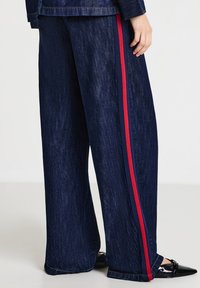 Pantaloni in denim a gamba larga di colore blu scuro con una striscia verticale rossa lungo il lato. Presentano una finitura testurizzata e una vita standard.