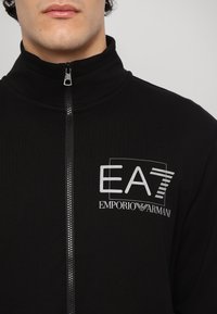 Black zip-up jacket with high collar, crafted from smooth fabric. Features a prominent EA7 logo and Emporio Armani branding on the chest.