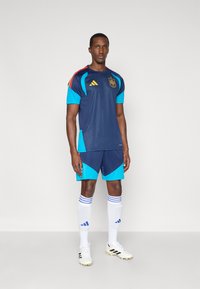 Vīriešu sportists stāv, ģērbies tumši zilā un tirkīza Spānijas futbola formā, baltās zeķēs un baltos sporta apavos, pozējot vienkāršā fonā.