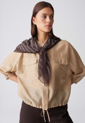 Woman with straight brown hair wearing a beige buttoned blouse with puffed sleeves and a dark striped scarf tied around her neck.