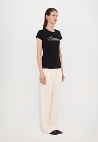 Schwarzes T-Shirt mit kurzen Ärmeln und metallic-"Armani Exchange"-Logo, kombiniert mit fließenden, cremefarbenen Hosen und schwarzen Slippern.
