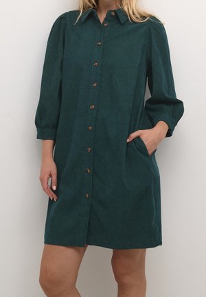 Woman wearing a dark green button-up dress with three-quarter sleeves, a collar, and side pockets, standing against a plain background.