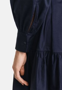 Rochie bleu marin cu un design în straturi, având mâneci puff lungi și mansete cu nasturi. Fabricată dintr-un material neted cu un ușor luciu.
