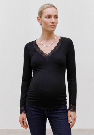 Donna incinta che indossa un top nero a maniche lunghe con rifiniture in pizzo e jeans scuri, guarda verso destra su uno sfondo semplice.