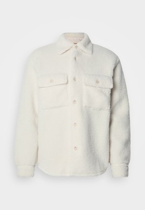 Casaco de camisa branco felpudo com dois bolsos no peito, fecho frontal com botões e colarinho clássico. Textura suave e acabamento em bainha arredondada.