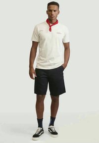 T-shirt creme de manga curta com um gráfico vermelho no peito, combinado com calções pretos e ténis de skate preto e branco. Banda vermelha à volta do pescoço.