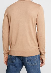 Lysebrun strikket sweater med lange ærmer, ribbede manchetter og kant, samt en glat tekstur, båret over blå denimjeans.