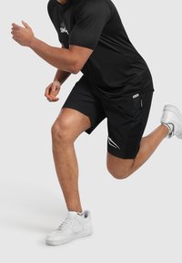Svarta sportshorts med sidofickor och en vit logotyp, ihop med en svart kortärmad skjorta och vita sneakers.
