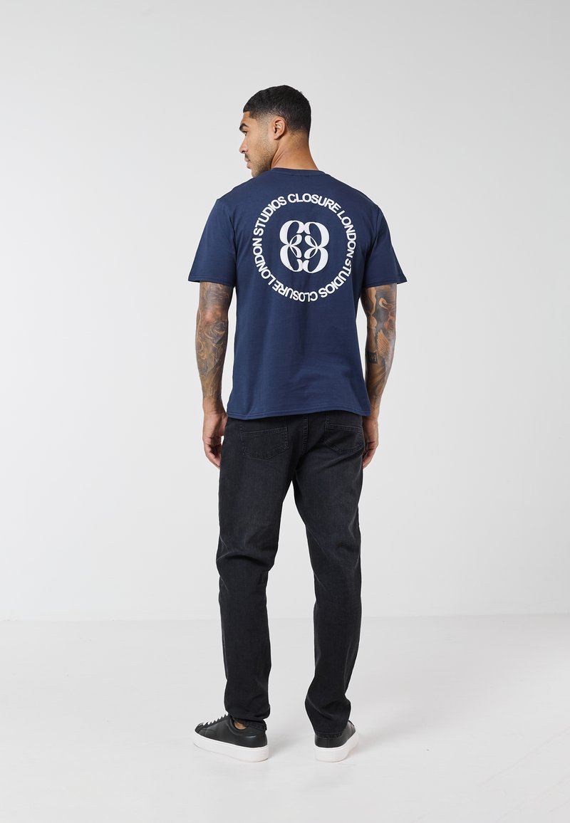 Male model with tattoos wearing a navy blue graphic T-shirt, black jeans, and black sneakers, standing against a plain white background.